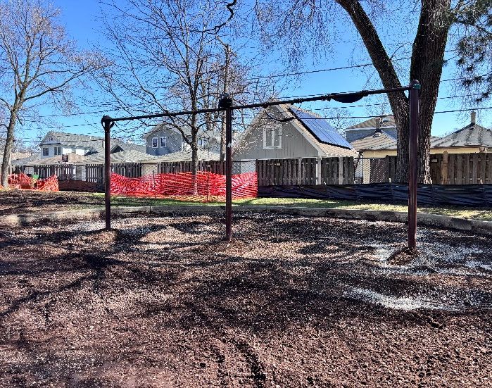 Andersen Park: A metal swing set frame stands on a mulched ground, with a red safety fence and houses in the background.