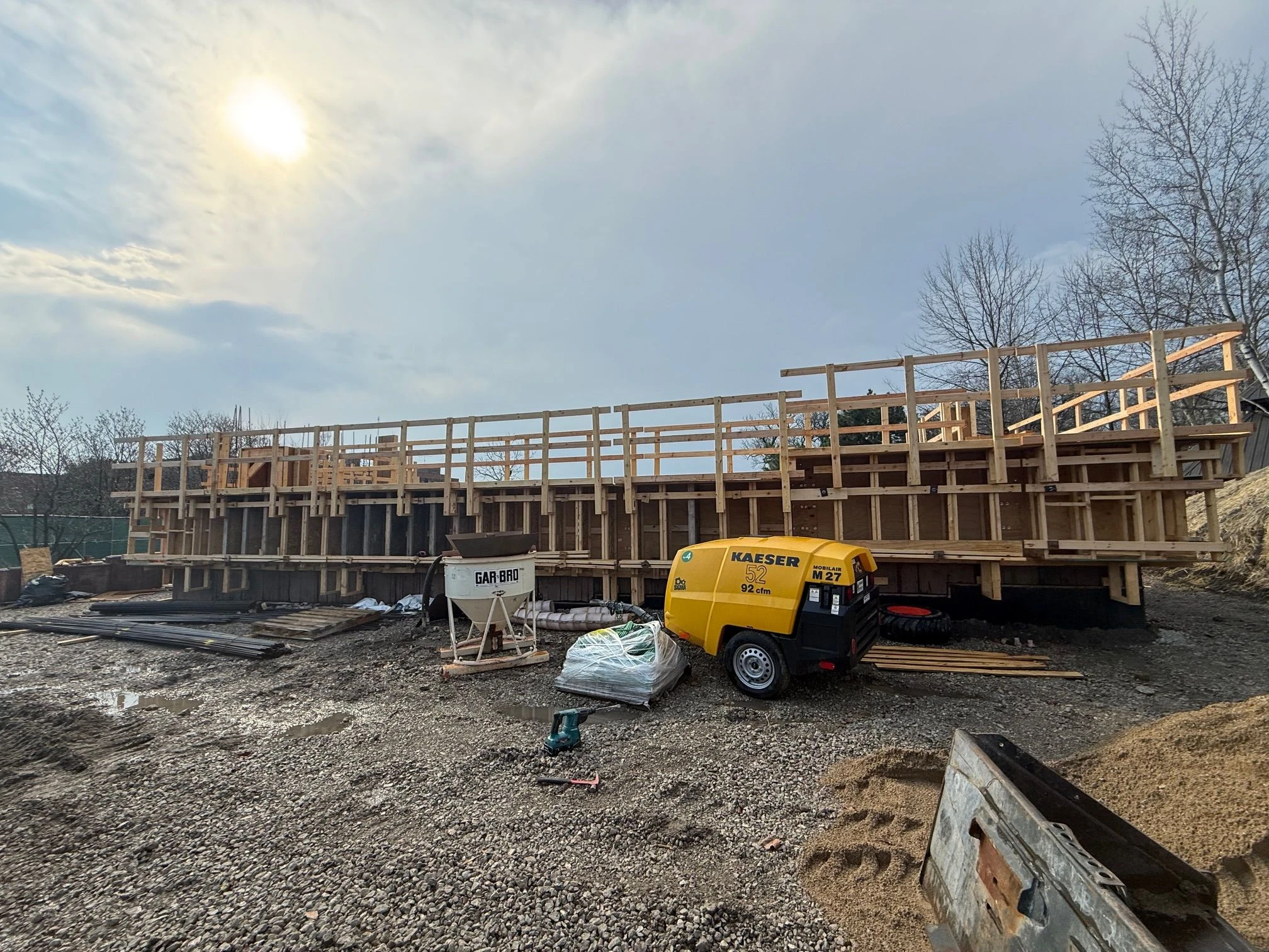 Construction site with wooden formwork for a concrete structure, a yellow Kaeser air compressor, and a Gar Bro concrete