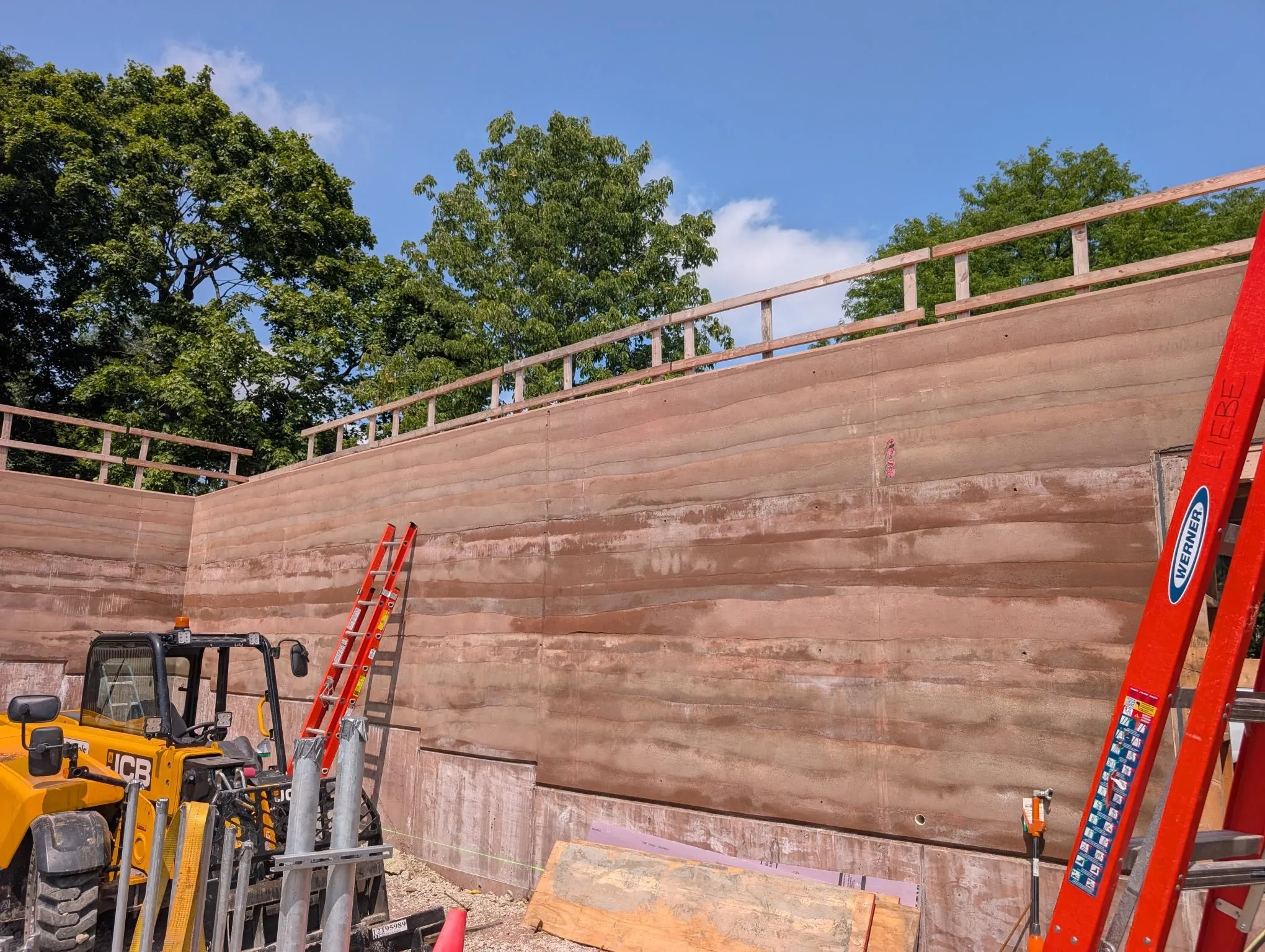 Construction of a rammed earth wall at Field Park, with a yellow JCB loader and red Werner ladders present.