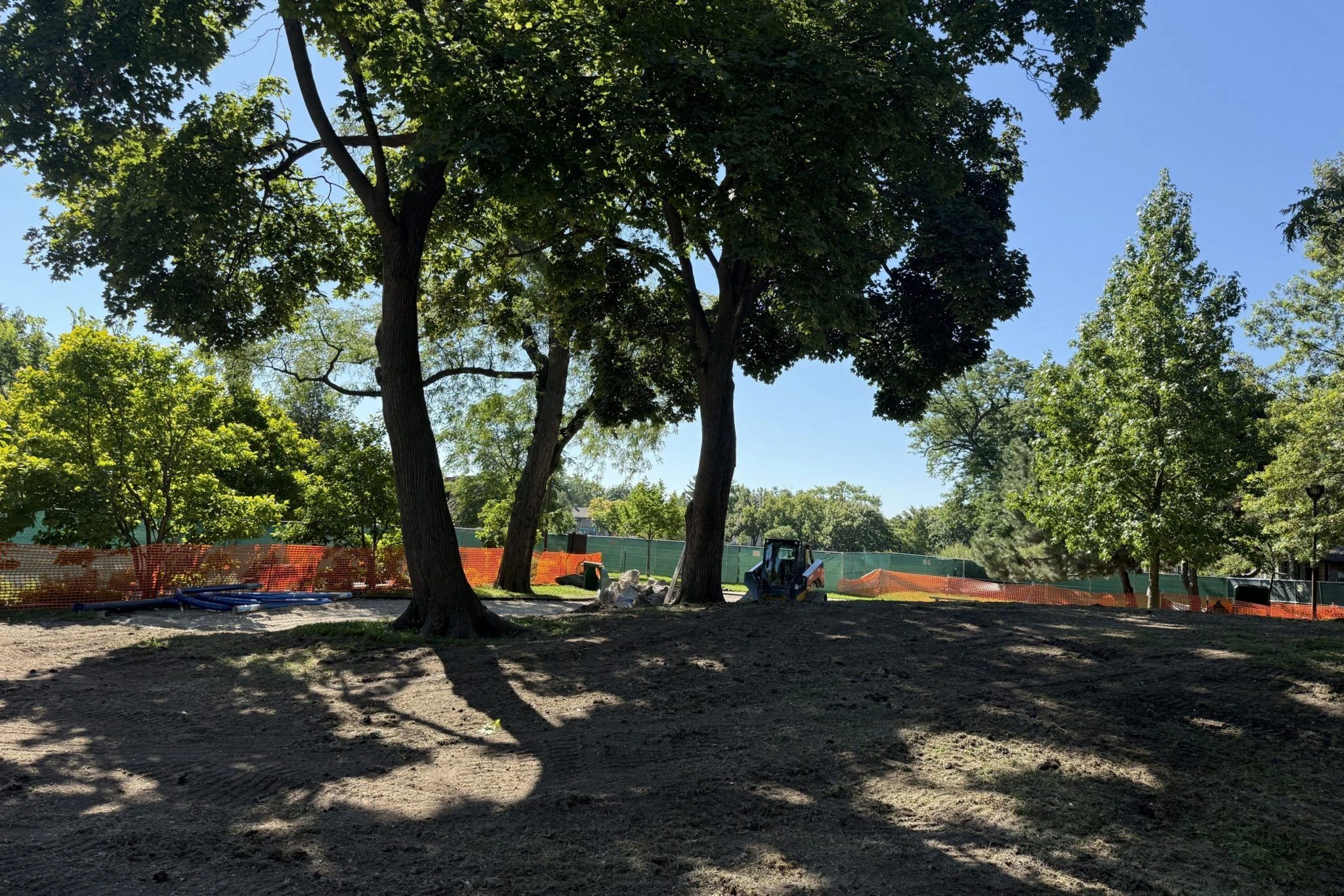 Construction equipment and materials are present in a park area undergoing landscaping, with orange safety fencing visible.