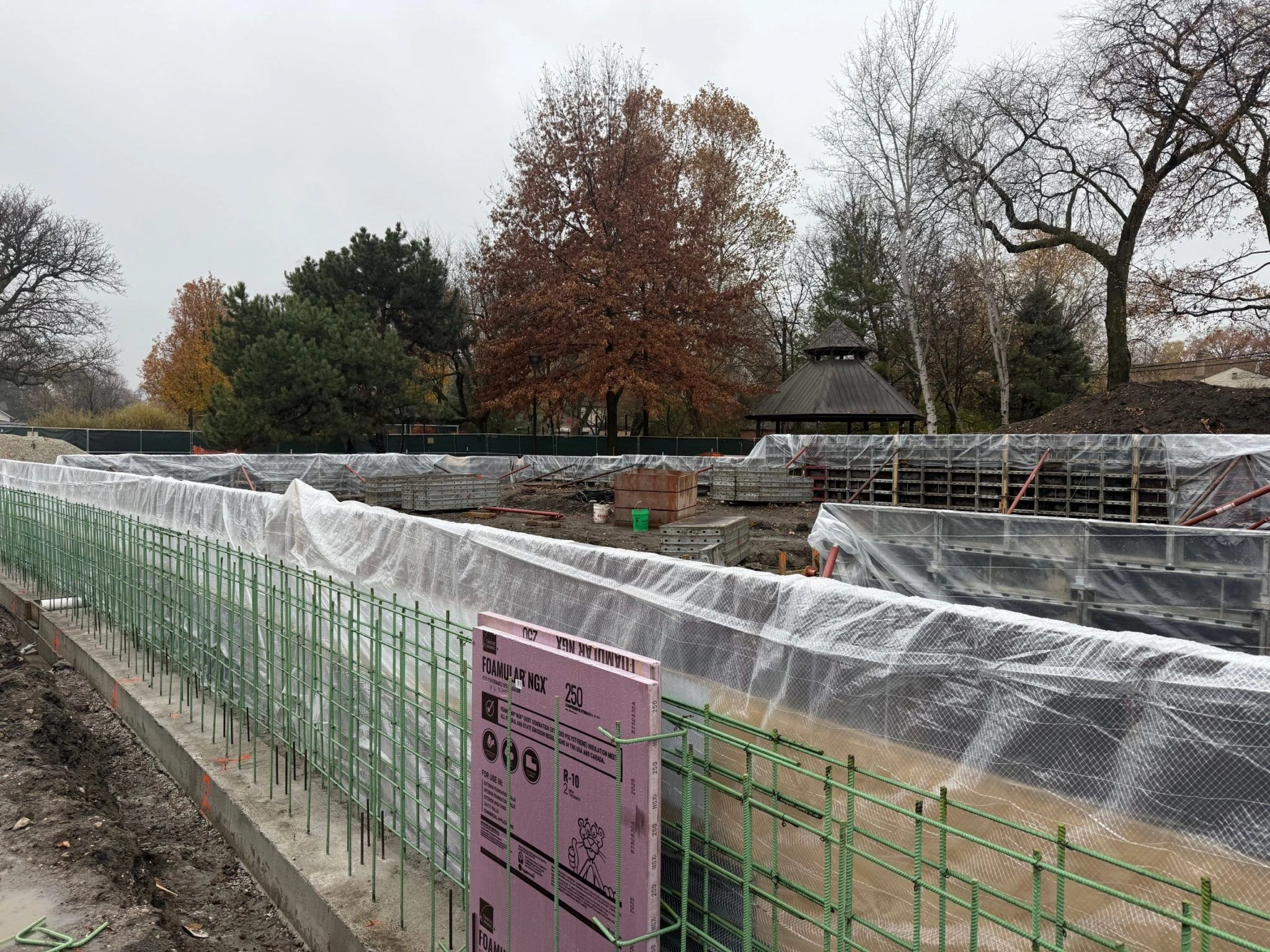 Construction site at Field Park with rebar and concrete forms covered in plastic sheeting. A gazebo is visible in the