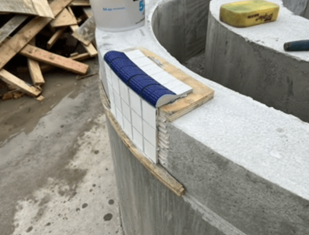 Construction of a curved pool edge with white tiles and blue mosaic tiles being installed.