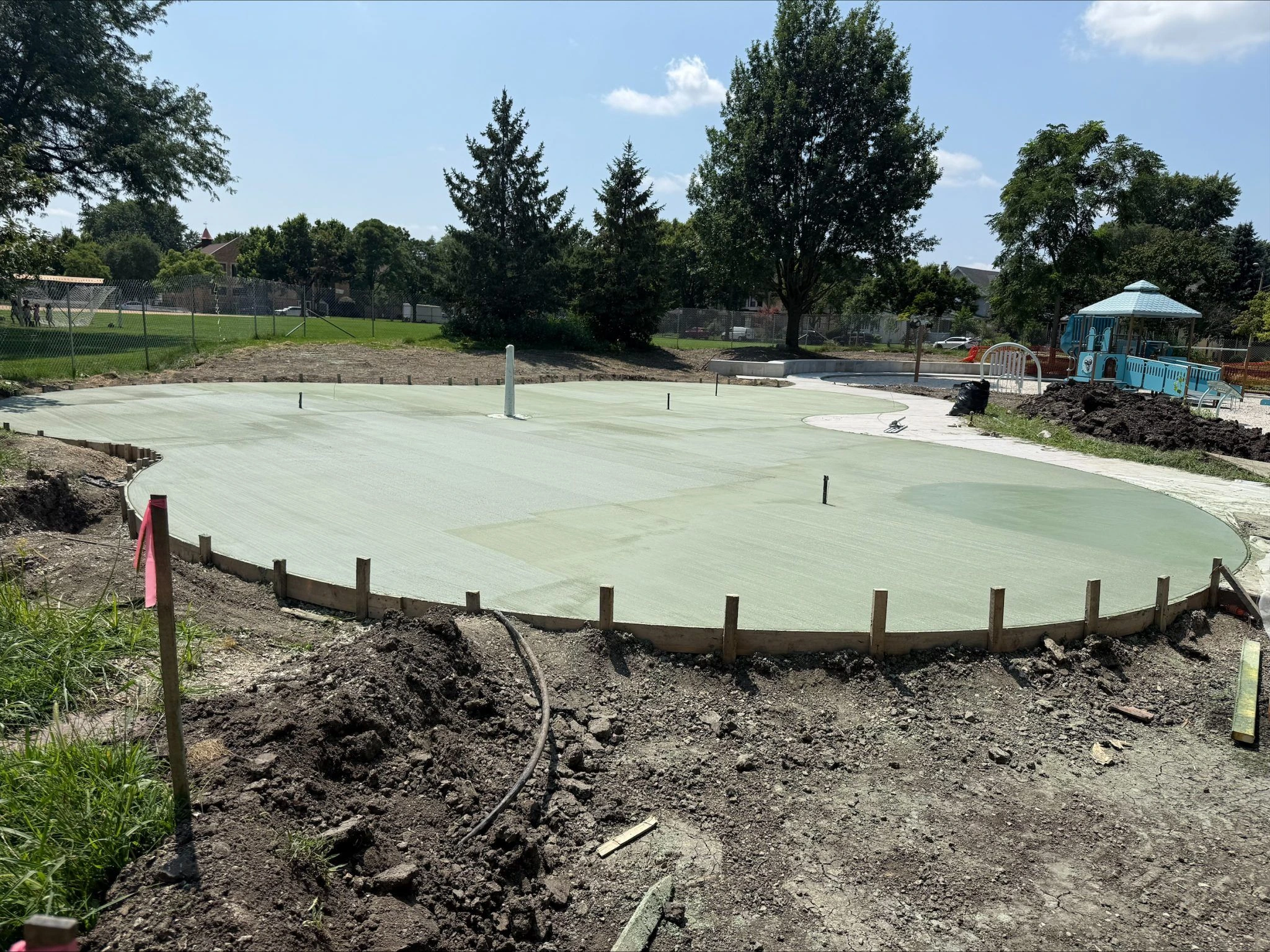 Newly poured concrete splash pad area at Longfellow Park, with surrounding construction materials and playground equipment