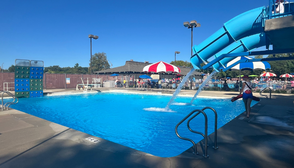 A lifeguard walks near a bright blue swimming pool with a water slide pouring water into it. A climbing wall is visible on