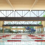 Interior view of a modern indoor aquatics facility with a swimming pool, lanes, a blue water slide, and people swimming and