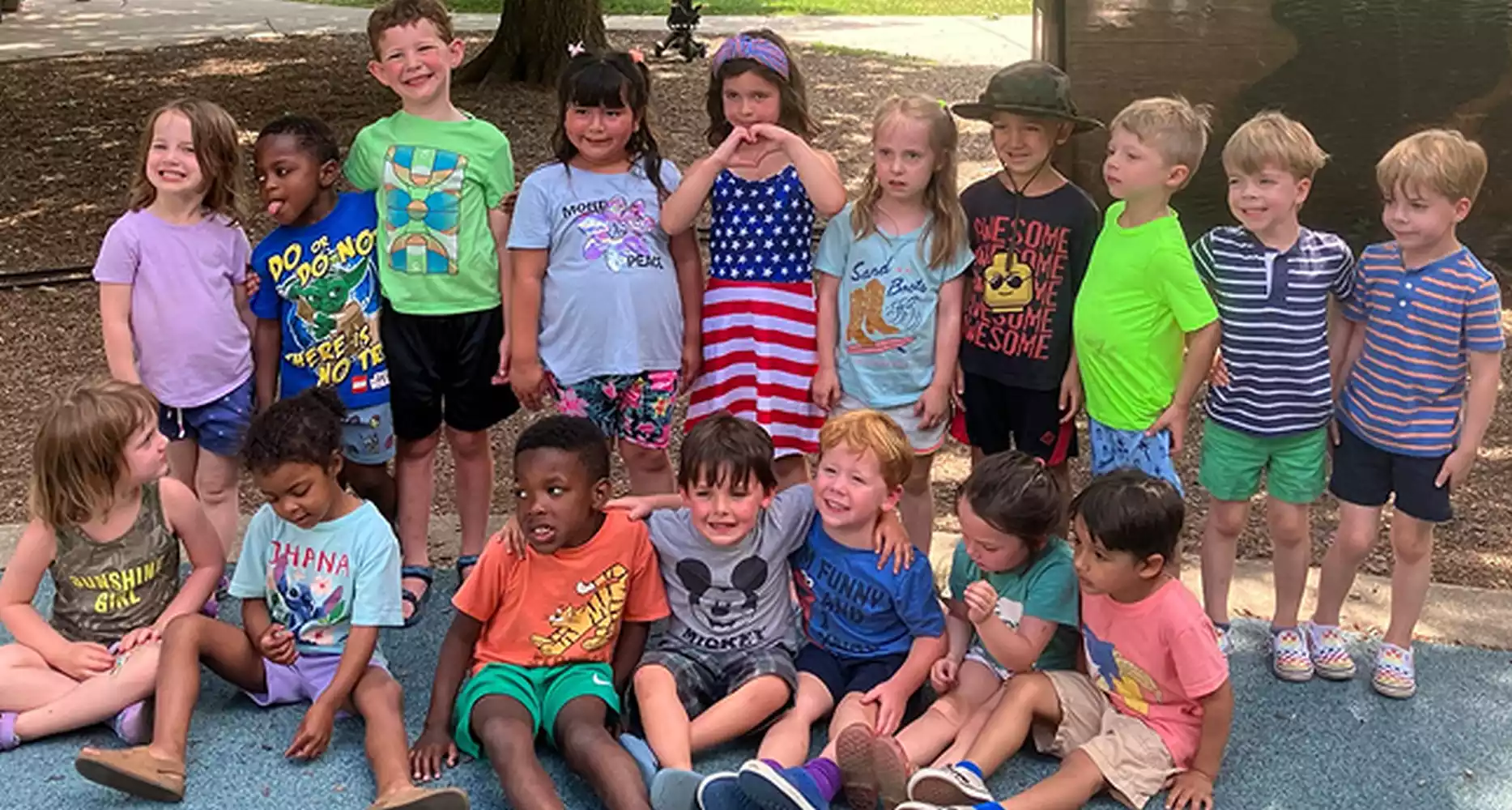 A diverse group of young children, some standing and some sitting, pose for a photo outdoors. Many are smiling.