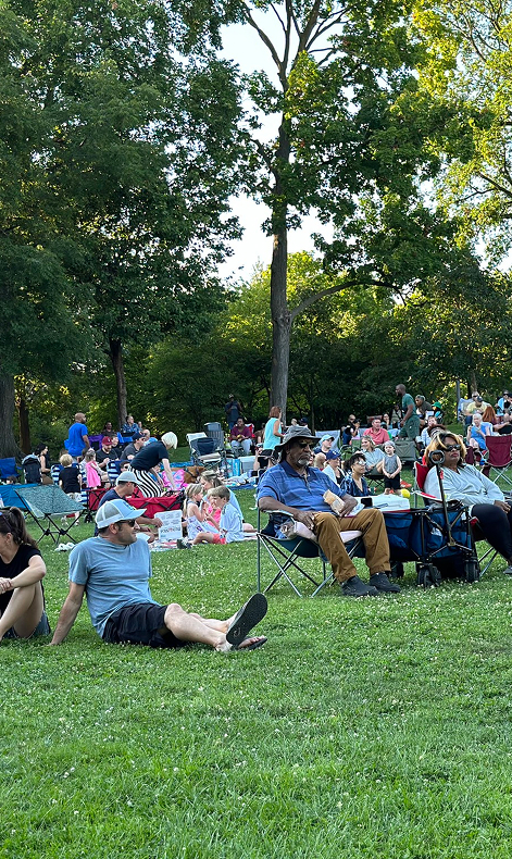 People relax on the grass in a park, seated in lawn chairs and on blankets, enjoying an outdoor event.