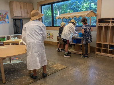 Children in straw hats and lab coats engage in an activity at a play station in a classroom.