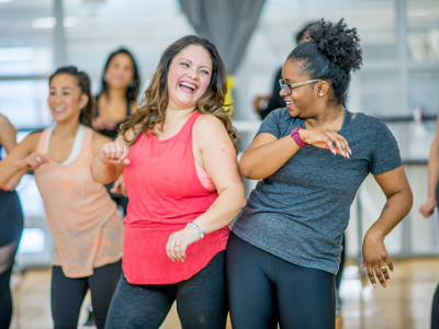 Two women laughing and dancing in a fitness class. They are in the foreground, with other participants blurred in the