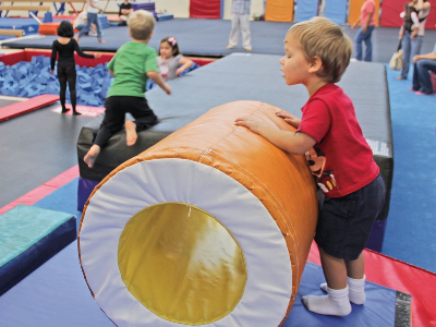 A young boy in a red shirt and dark shorts interacts with a large orange cylindrical mat in a gymnastics gym.