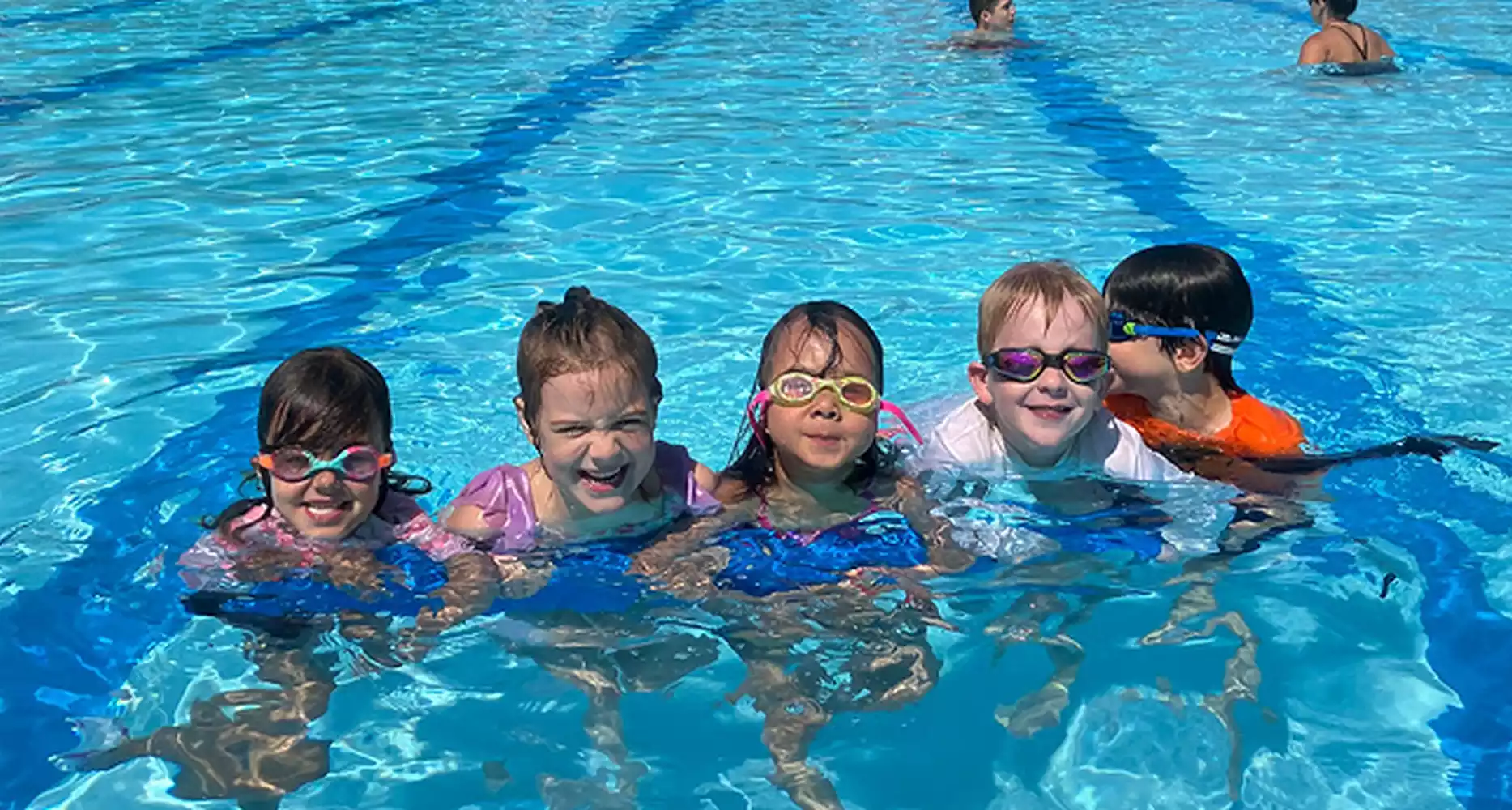 Five children wearing goggles smile as they float in a swimming pool.