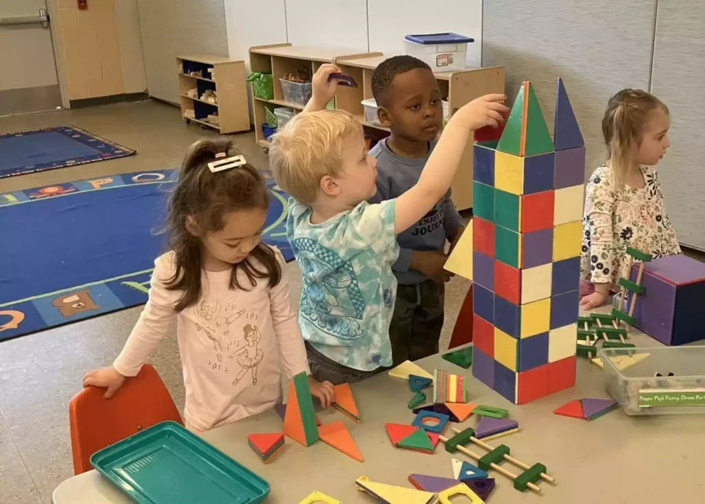 Four young children collaborate to build a colorful tower with magnetic blocks in a classroom setting.