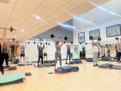 Group of people performing BODYPUMP class, lifting barbells overhead in a fitness studio.