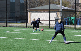 Three people play soccer on an artificial turf field with white markings. One person in the foreground faces away.