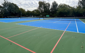 Outdoor tennis courts with blue playing surfaces and green surrounds, set against a backdrop of autumn trees. A small inset