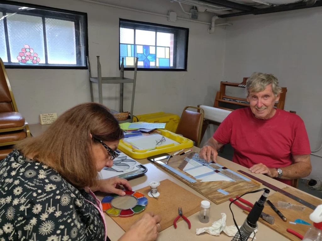 A woman and a man work on stained glass projects at a table. The woman is focused on a circular piece, while the man smiles