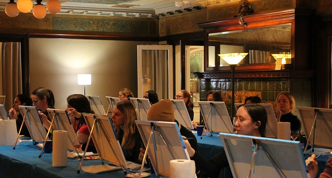 Adults gathered around a table, focused on painting canvases on easels in a dimly lit room.