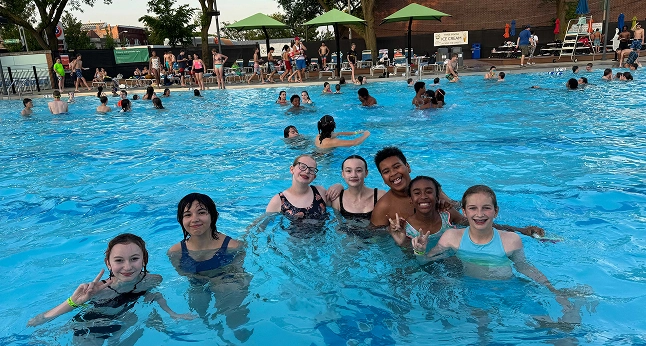 A group of diverse children and teens smile and pose for a photo in a crowded outdoor swimming pool.