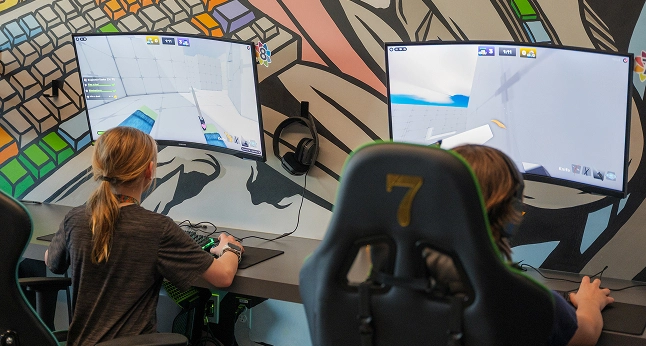 Two children play video games on curved monitors in a room with colorful, abstract wall art.