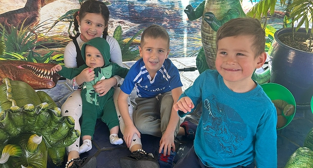 Four children posing in front of a dinosaur-themed backdrop. A baby in a green dinosaur outfit is held by a girl.