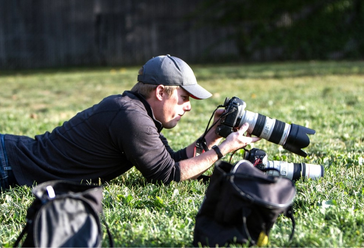 Photographer lying on grass, holding a professional camera with a long lens, with another camera and bags nearby.
