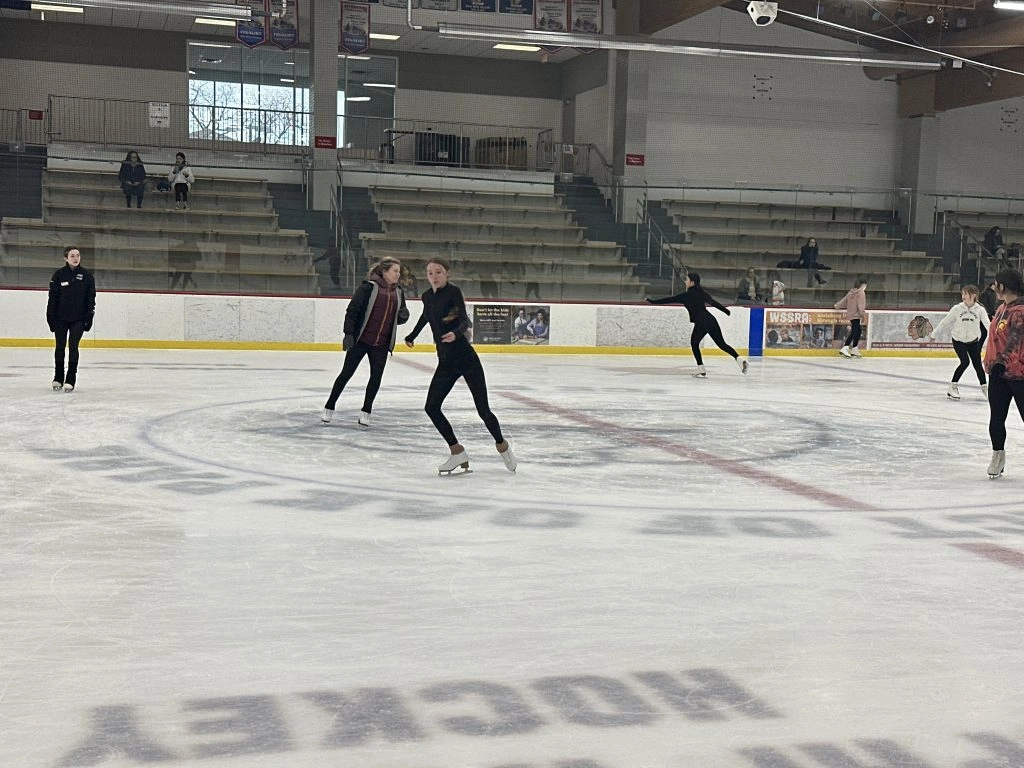 Several figure skaters practice on an ice rink with bleachers in the background.
