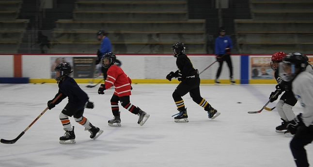 Young hockey players in uniform skate on an ice rink during practice, with coaches in the background.