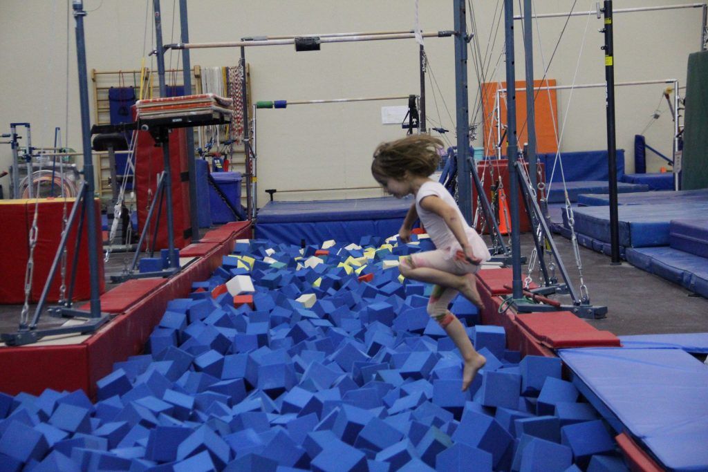 A young girl in athletic wear jumps into a pit filled with blue foam cubes in a gymnastics gym.