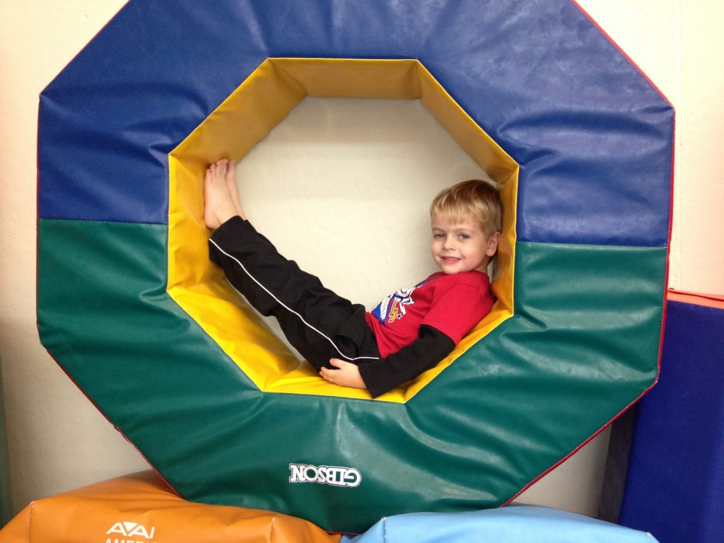 A young boy smiles while sitting inside a colorful octagonal gymnastics tunnel.