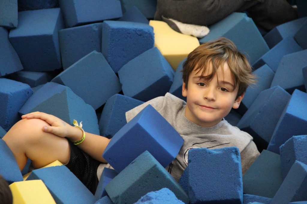 A young boy with brown hair smiles while lying in a pit of blue foam cubes.