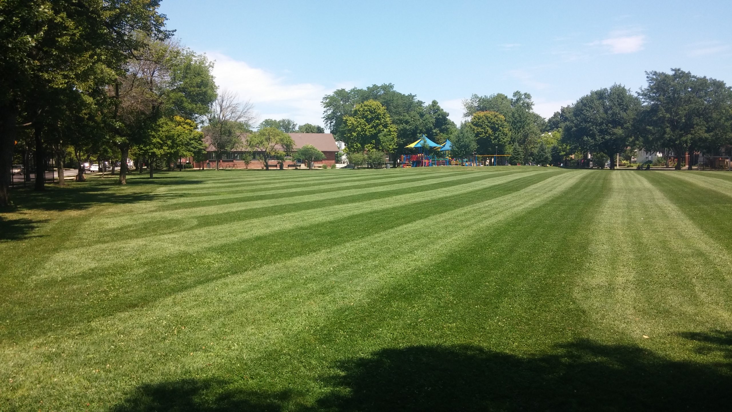 A wide, freshly mown lawn with striped patterns leads towards a playground and buildings under a clear blue sky.
