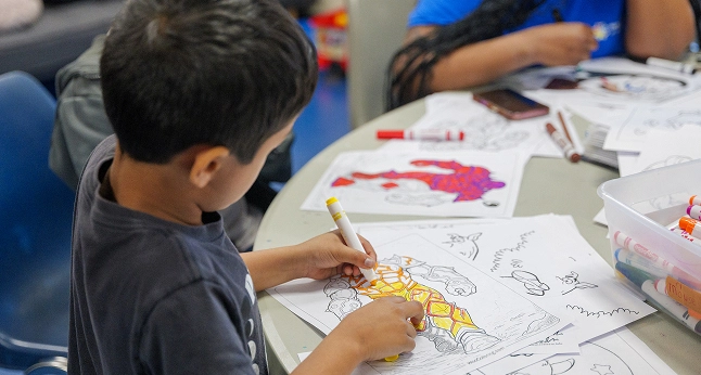 A young boy colors a superhero drawing with a yellow marker at a table with other children and art supplies.