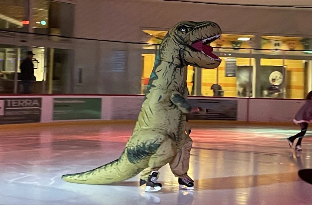 A person in a T-Rex costume ice skating on an indoor rink.
