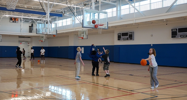 Several teenagers play basketball in a gymnasium. A clock on the wall shows 5:12.