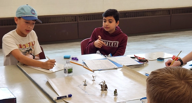 Two teens at a table are engaged in a tabletop role-playing game, with one drawing and the other gesturing.
