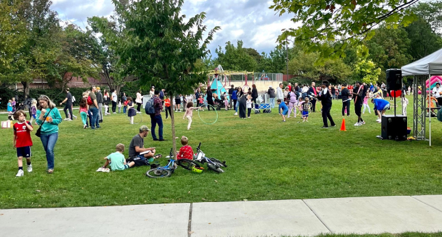 A diverse group of people, including children and adults, enjoy an outdoor event on a grassy field with a playground in the