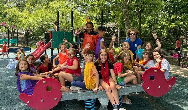 A group of children and a counselor pose for a photo on a playground seesaw during a summer camp activity.