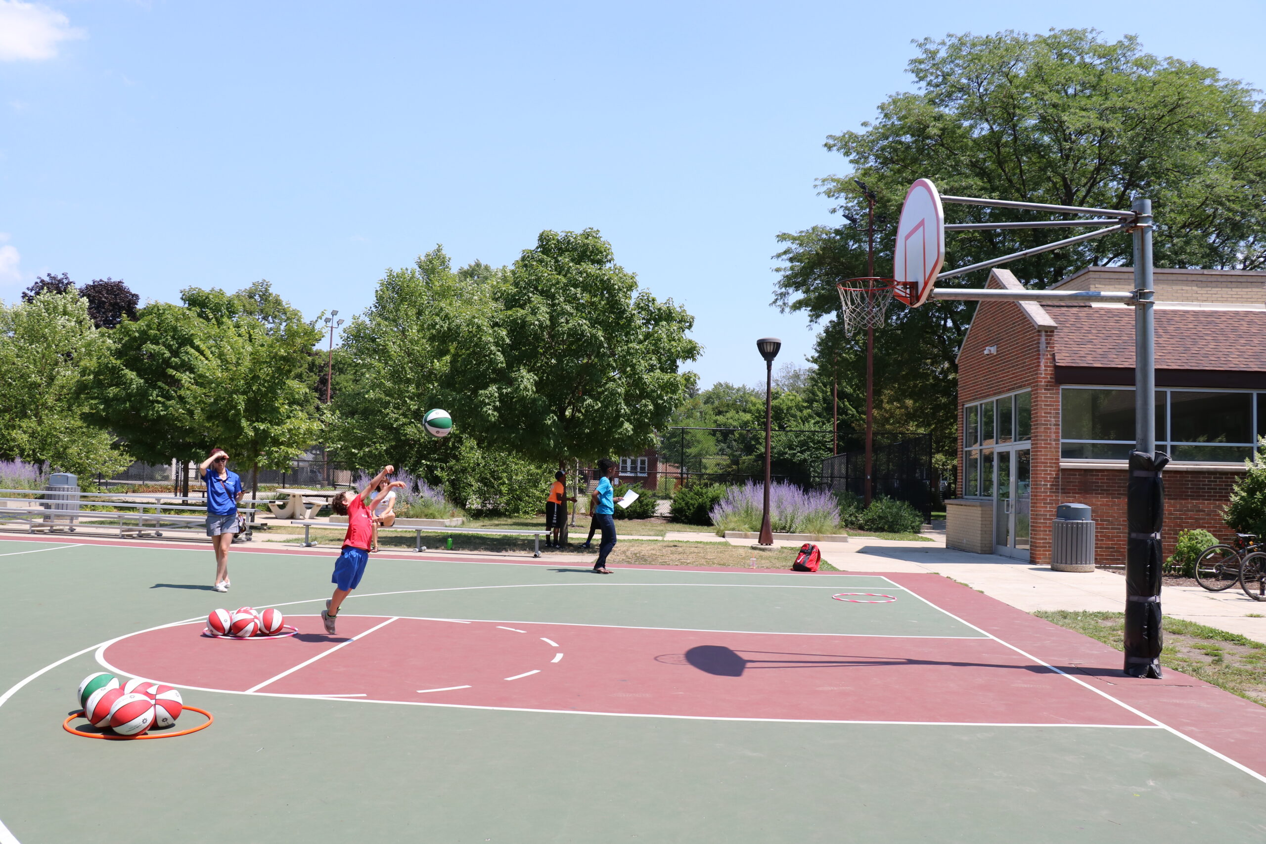 A young person in a red shirt jumps to shoot a basketball on an outdoor court. Other people and basketballs are visible.