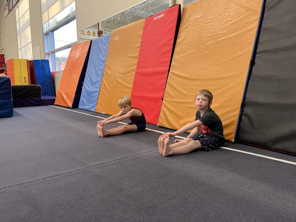 Two young boys stretch on a gymnastics mat in front of colorful mats. One boy wears a leotard, the other a t-shirt and