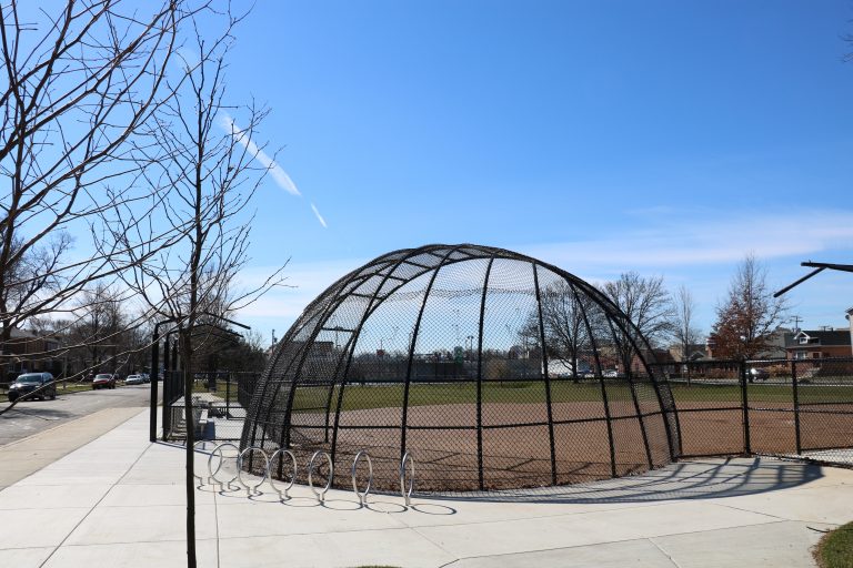 A domed batting cage with a chain-link fence encloses a baseball field under a clear blue sky.