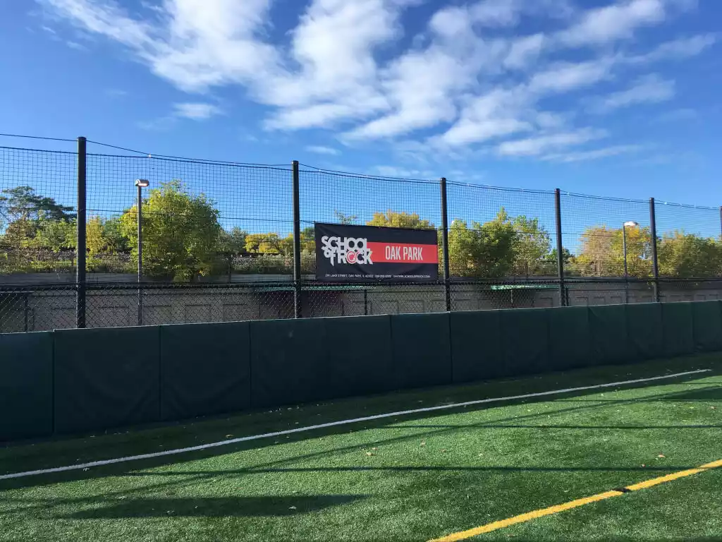 A banner for School of Rock Oak Park hangs on a fence above a green turf field.