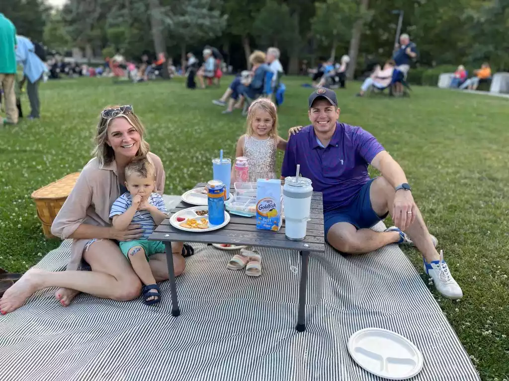 A family of four enjoys a picnic on a striped blanket in a park with other people in the background.