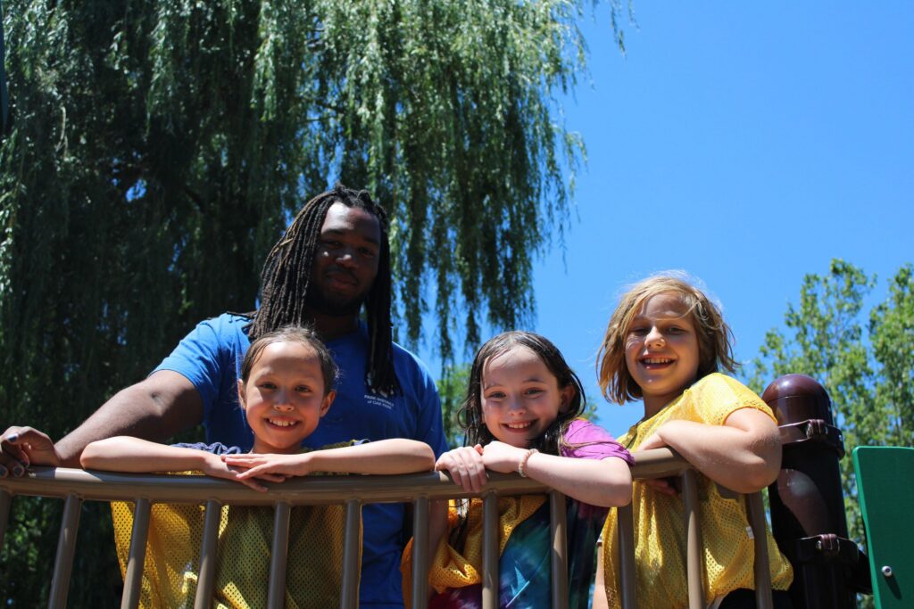 A counselor with dreadlocks stands behind three smiling children on a playground structure.