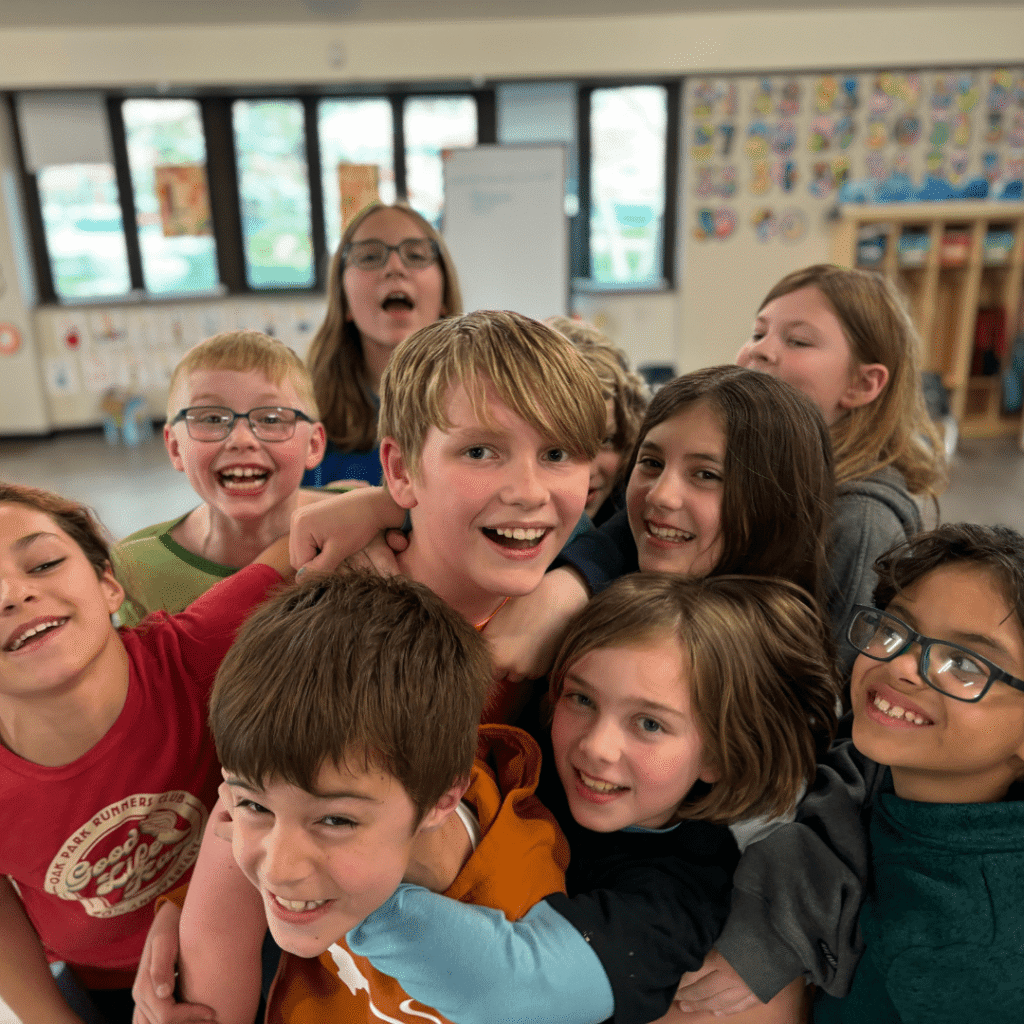 A diverse group of smiling children in a classroom setting, huddled together and looking at the camera.