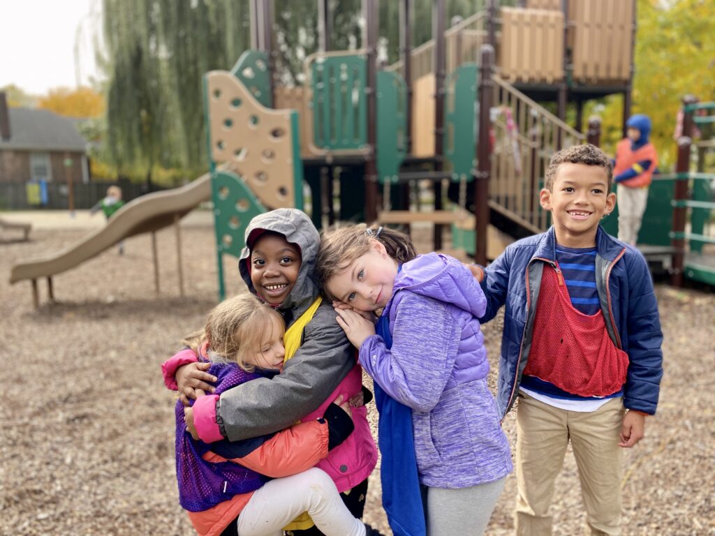 Four children hugging on a playground with a slide and play structure in the background.