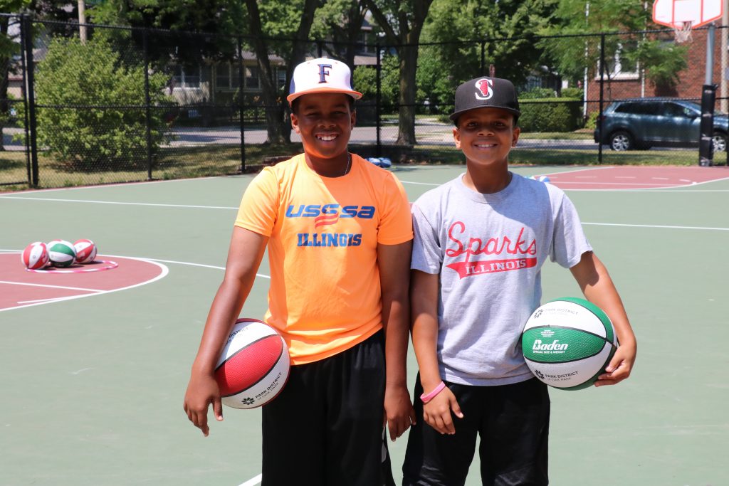 Two young boys stand on a basketball court holding basketballs. The boy on the left wears an orange shirt and the boy on the