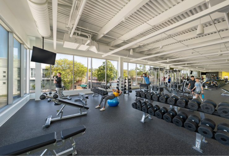 A bright, modern fitness center with large windows, weight racks, exercise machines, and people working out.