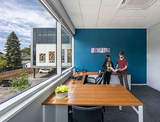 Two people stand near a desk in a modern office with a large window and a teal accent wall that says "INSPIRE".