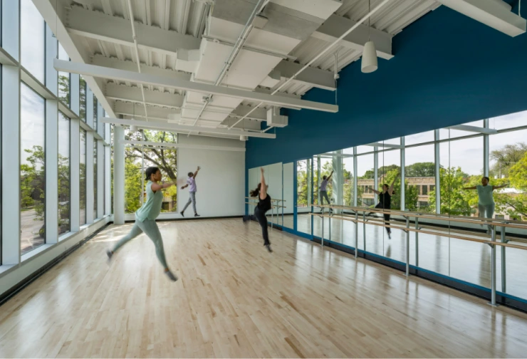 Dancers practice in a bright studio with large windows, mirrors, and ballet barres.