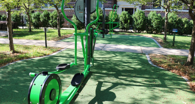 A bright green elliptical machine for outdoor fitness is set up on a green rubberized surface with a walking path.